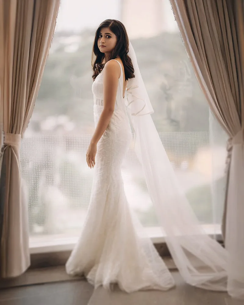 Bride in a white gown with a long veil standing by a window.