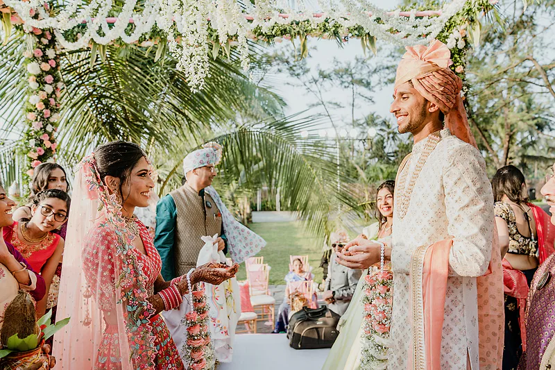 Bride in pink lehenga and groom in sherwani exchanging vows outdoors.
