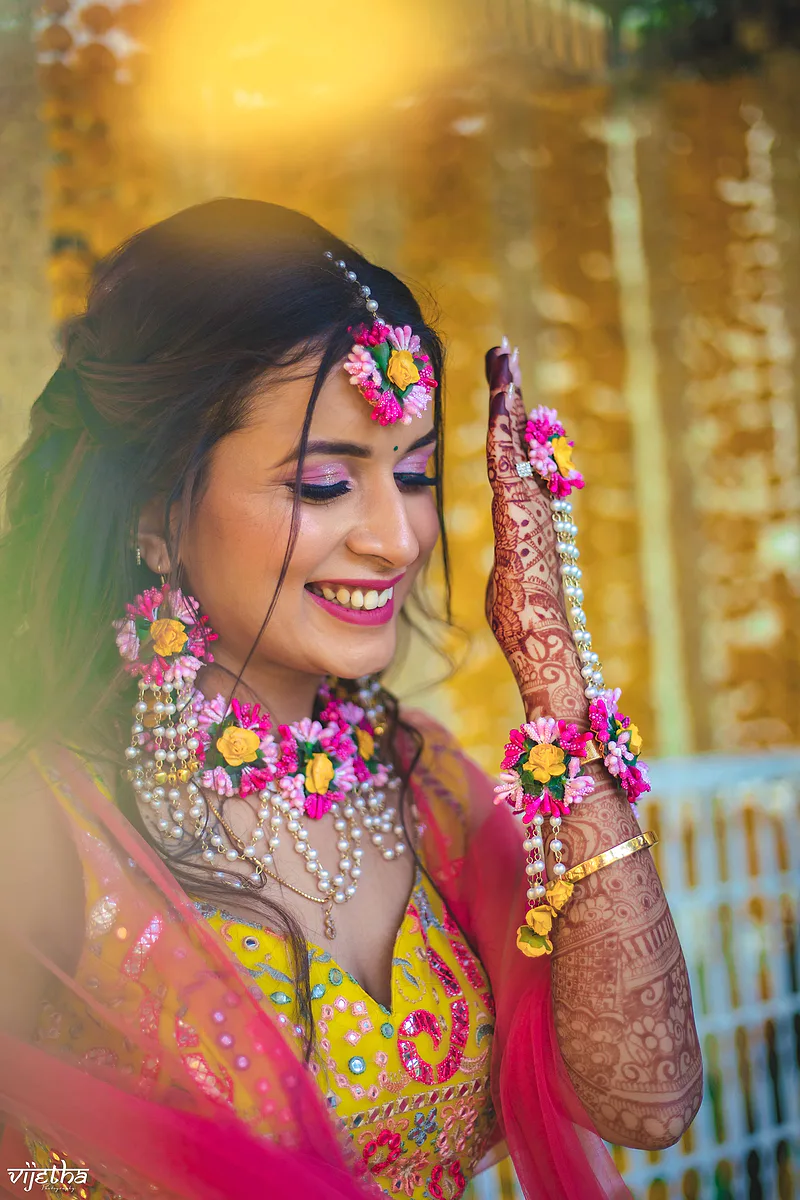 Bride with floral jewelry, intricate mehendi, and a vibrant yellow and pink outfit.